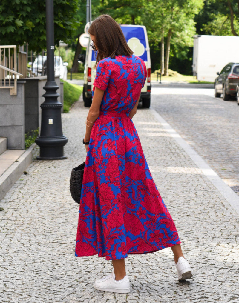 Red - A woman walking on a cobblestone street wearing a vibrant pink and green floral print midi dress with a V-neckline, short sleeves, a tie-waist, and a front slit.