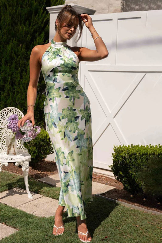A woman in a sunny garden wearing a full-length, satin-finish halter dress with a green floral print and a gathered high neckline.