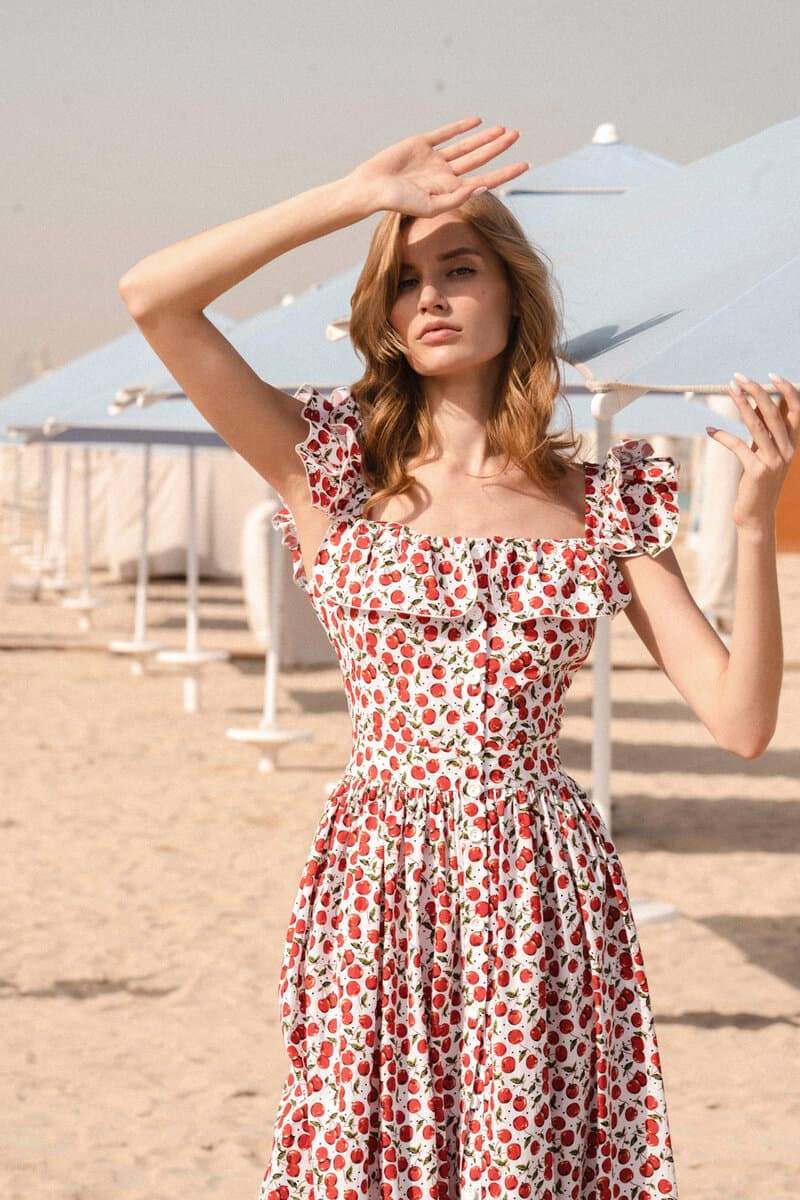 A woman wears a white fit-and-flare midi dress covered in a red cherry print, featuring a square neckline, ruffled cap sleeves, a fitted bodice, and a full skirt with a ruffled hem.