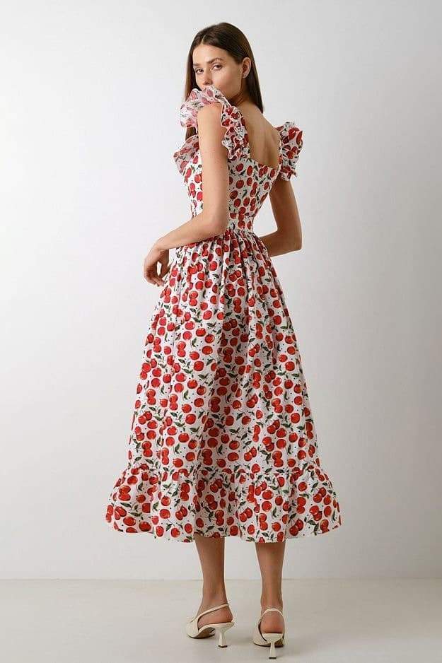 A woman wears a white fit-and-flare midi dress covered in a red cherry print, featuring a square neckline, ruffled cap sleeves, a fitted bodice, and a full skirt with a ruffled hem.