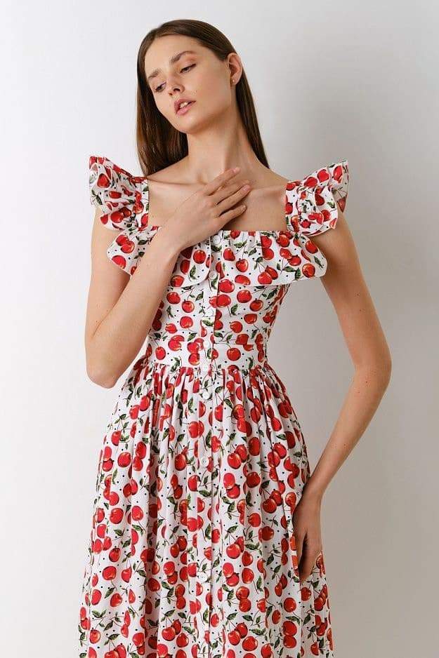 A woman wears a white fit-and-flare midi dress covered in a red cherry print, featuring a square neckline, ruffled cap sleeves, a fitted bodice, and a full skirt with a ruffled hem.