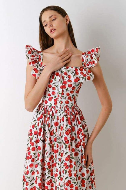 A woman wears a white fit-and-flare midi dress covered in a red cherry print, featuring a square neckline, ruffled cap sleeves, a fitted bodice, and a full skirt with a ruffled hem.