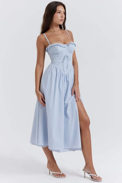 A woman stands against a white background wearing a light blue midi dress with thin spaghetti straps, a fitted eyelet corset bodice with a ruffled neckline, and a flowing A-line skirt with a high thigh slit.