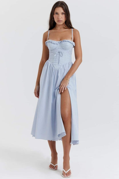 A woman stands against a white background wearing a light blue midi dress with thin spaghetti straps, a fitted eyelet corset bodice with a ruffled neckline, and a flowing A-line skirt with a high thigh slit.