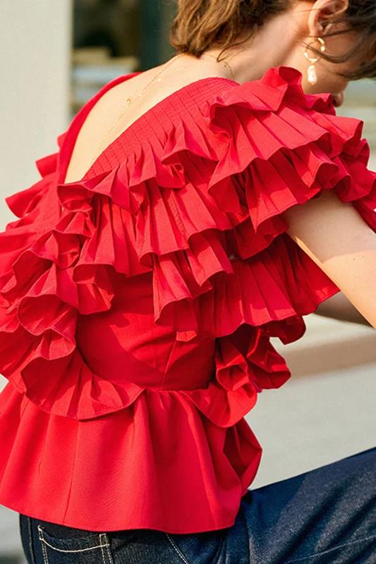 A blonde woman modelling a bright red peplum blouse with dramatic, voluminous ruffles framing a V-neckline, paired with light wash jeans.