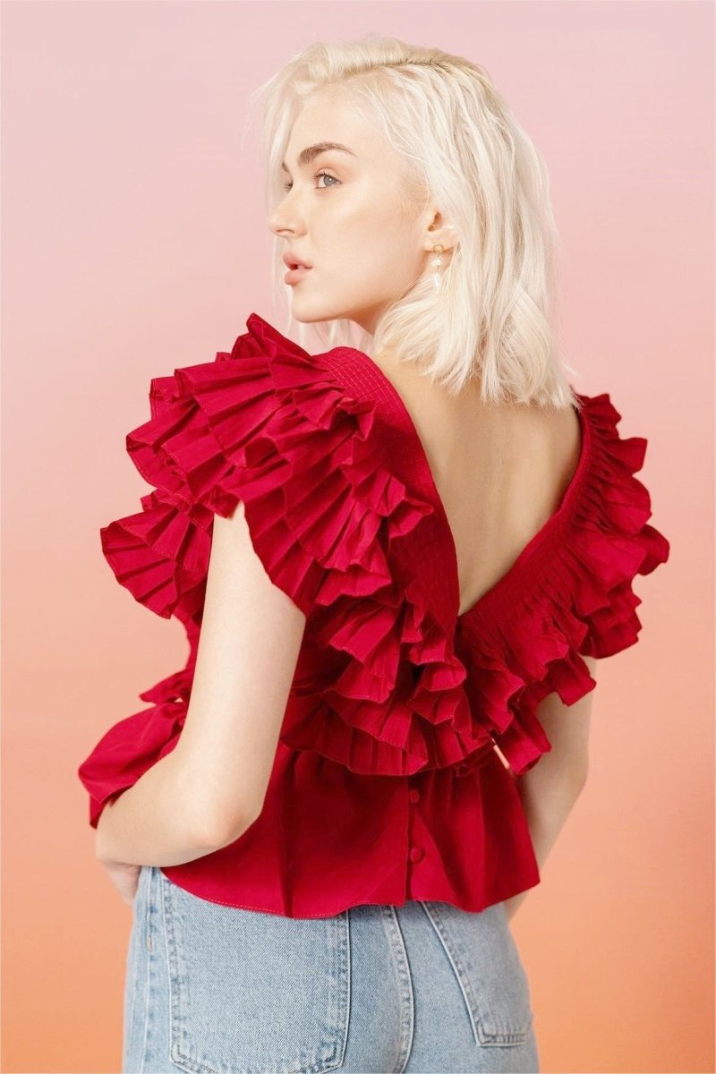 A blonde woman modelling a bright red peplum blouse with dramatic, voluminous ruffles framing a V-neckline, paired with light wash jeans.