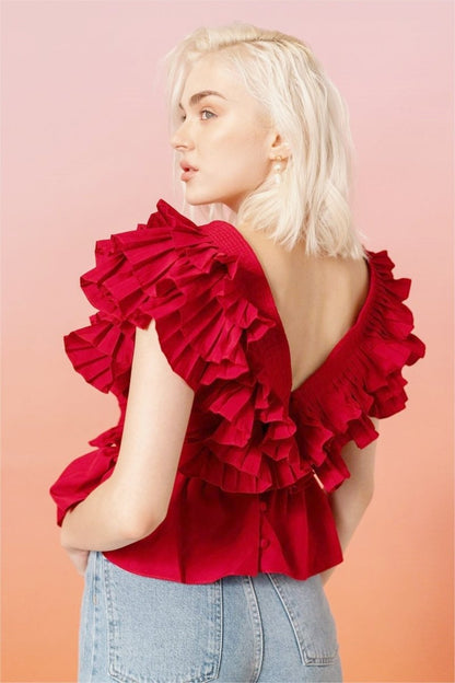 A blonde woman modelling a bright red peplum blouse with dramatic, voluminous ruffles framing a V-neckline, paired with light wash jeans.
