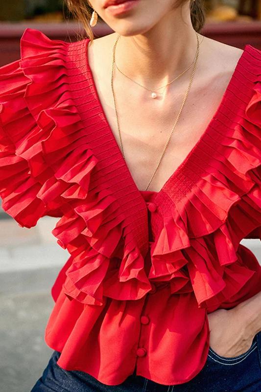 A blonde woman modelling a bright red peplum blouse with dramatic, voluminous ruffles framing a V-neckline, paired with light wash jeans.