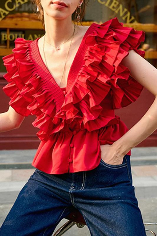 A blonde woman modelling a bright red peplum blouse with dramatic, voluminous ruffles framing a V-neckline, paired with light wash jeans.