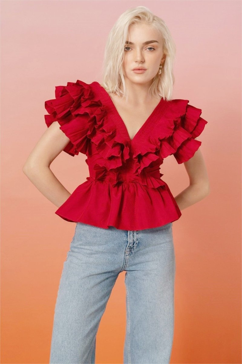 A blonde woman modelling a bright red peplum blouse with dramatic, voluminous ruffles framing a V-neckline, paired with light wash jeans.