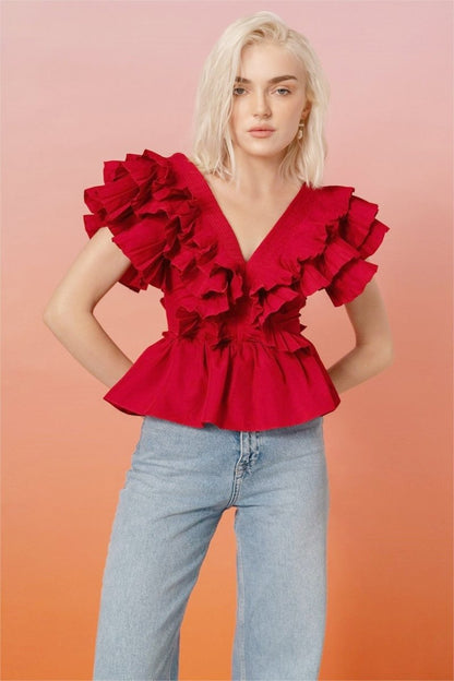 A blonde woman modelling a bright red peplum blouse with dramatic, voluminous ruffles framing a V-neckline, paired with light wash jeans.