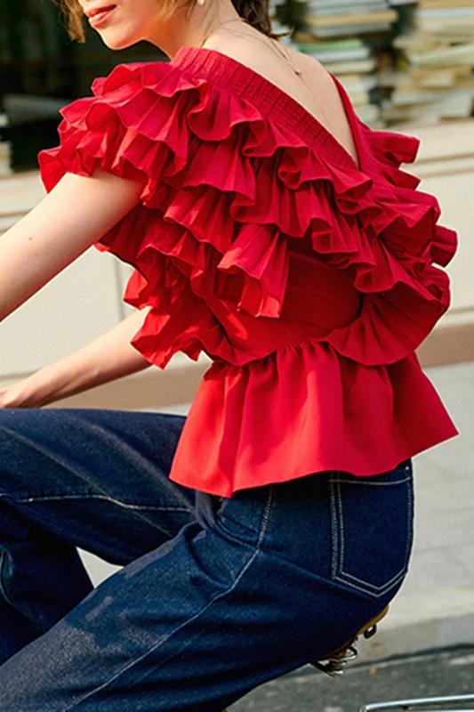 A blonde woman modelling a bright red peplum blouse with dramatic, voluminous ruffles framing a V-neckline, paired with light wash jeans.