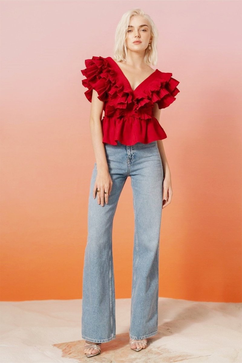 A blonde woman modelling a bright red peplum blouse with dramatic, voluminous ruffles framing a V-neckline, paired with light wash jeans.
