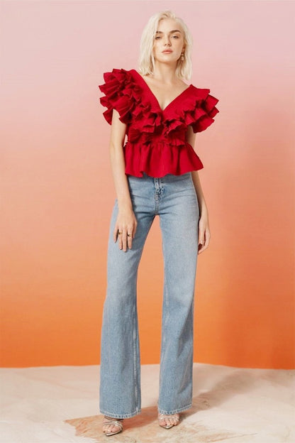 A blonde woman modelling a bright red peplum blouse with dramatic, voluminous ruffles framing a V-neckline, paired with light wash jeans.
