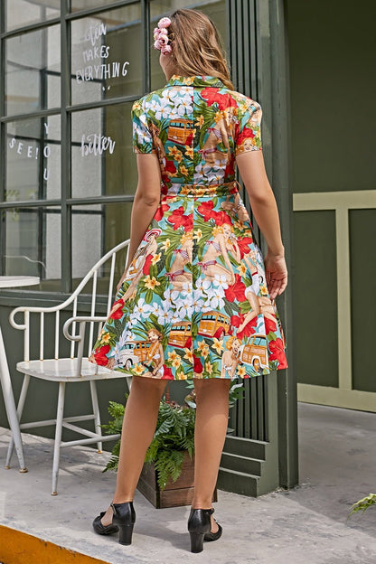 A woman with wavy brown hair and a pink flower clip smiles while wearing a short-sleeved, collared fit-and-flare dress. The dress features a colorful vintage tropical print with pin-up girls, hibiscus flowers, and woody wagons, and is cinched at the waist with a matching fabric belt.