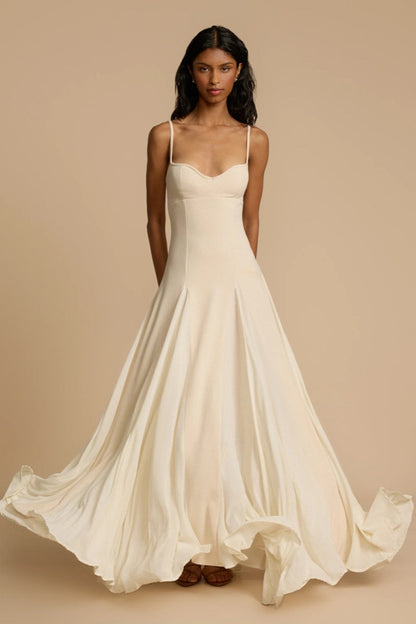 Full-length studio shot of a woman wearing a cream-colored A-line maxi dress with spaghetti straps, a sweetheart bustier bodice, and a voluminous, flowing skirt spread out at the hem.