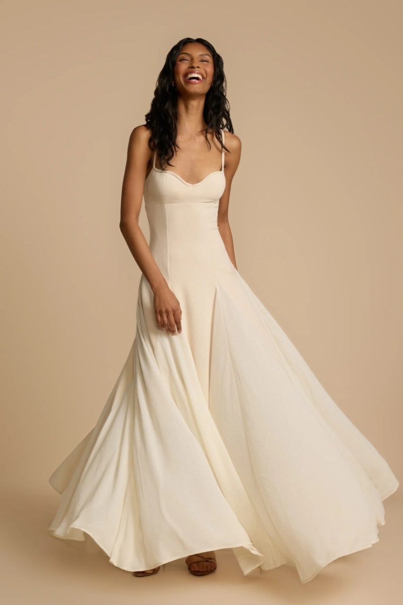 Full-length studio shot of a woman wearing a cream-colored A-line maxi dress with spaghetti straps, a sweetheart bustier bodice, and a voluminous, flowing skirt spread out at the hem.