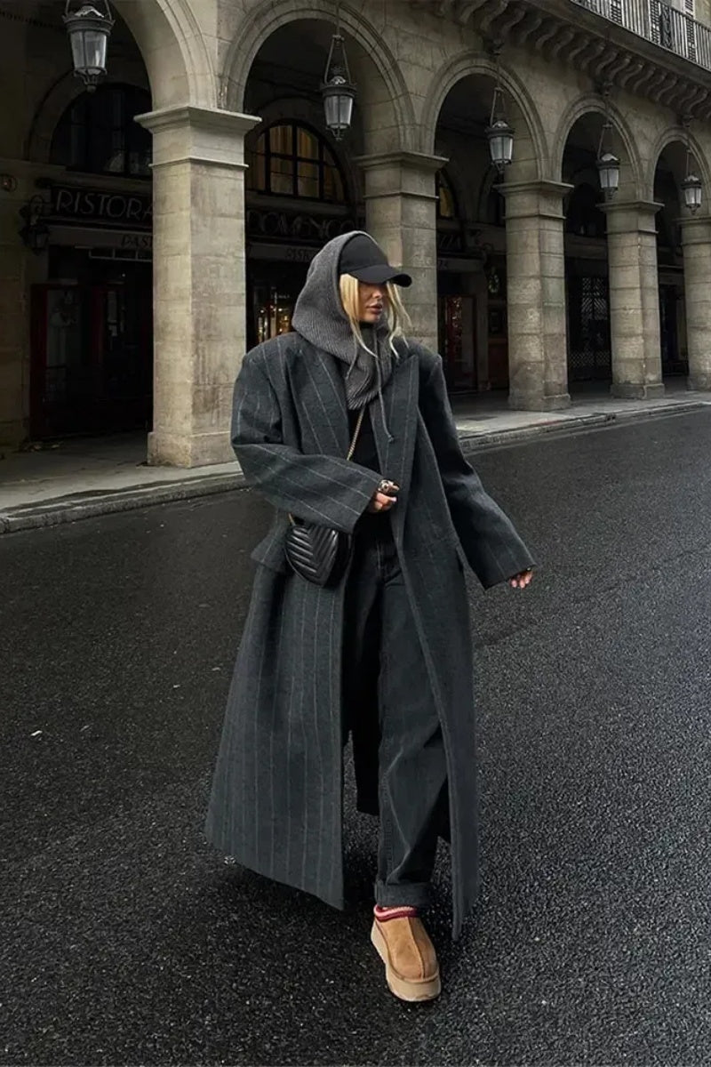 A woman walking on a city street wearing a dark grey, oversized maxi-length coat with subtle pinstripes, styled over dark jeans and a grey knit hood.
