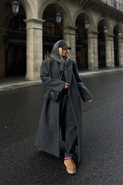 A woman walking on a city street wearing a dark grey, oversized maxi-length coat with subtle pinstripes, styled over dark jeans and a grey knit hood.