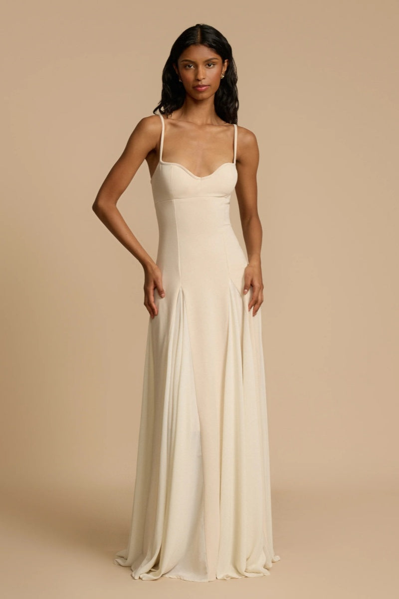 Full-length studio shot of a woman wearing a cream-colored A-line maxi dress with spaghetti straps, a sweetheart bustier bodice, and a voluminous, flowing skirt spread out at the hem.