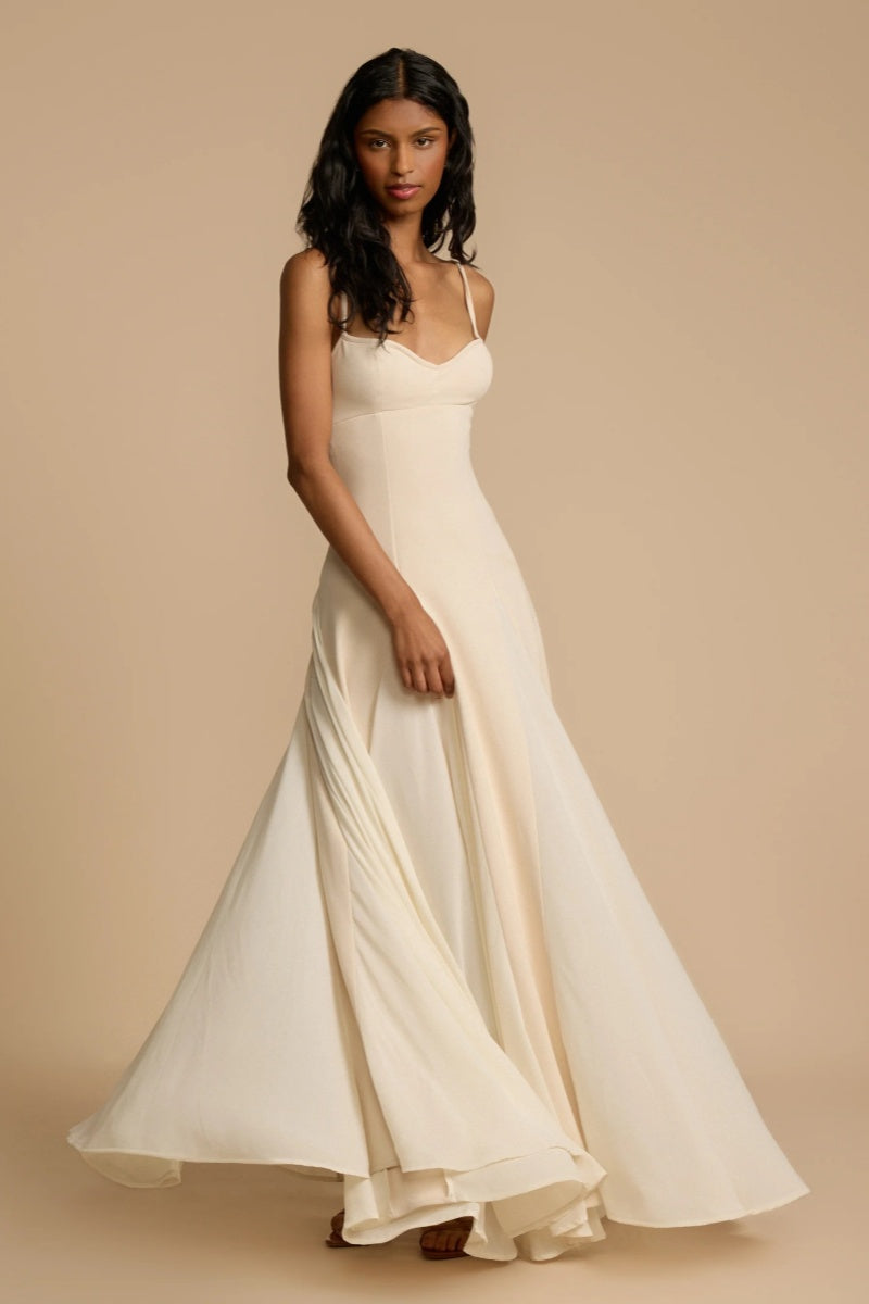 Full-length studio shot of a woman wearing a cream-colored A-line maxi dress with spaghetti straps, a sweetheart bustier bodice, and a voluminous, flowing skirt spread out at the hem.