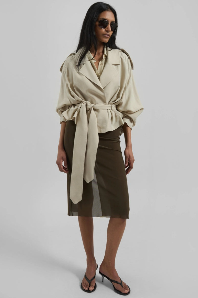 A woman models a light beige, cropped wrap trench jacket featuring wide lapels, shoulder epaulettes, belted cuffs, and a cinched waist tie.