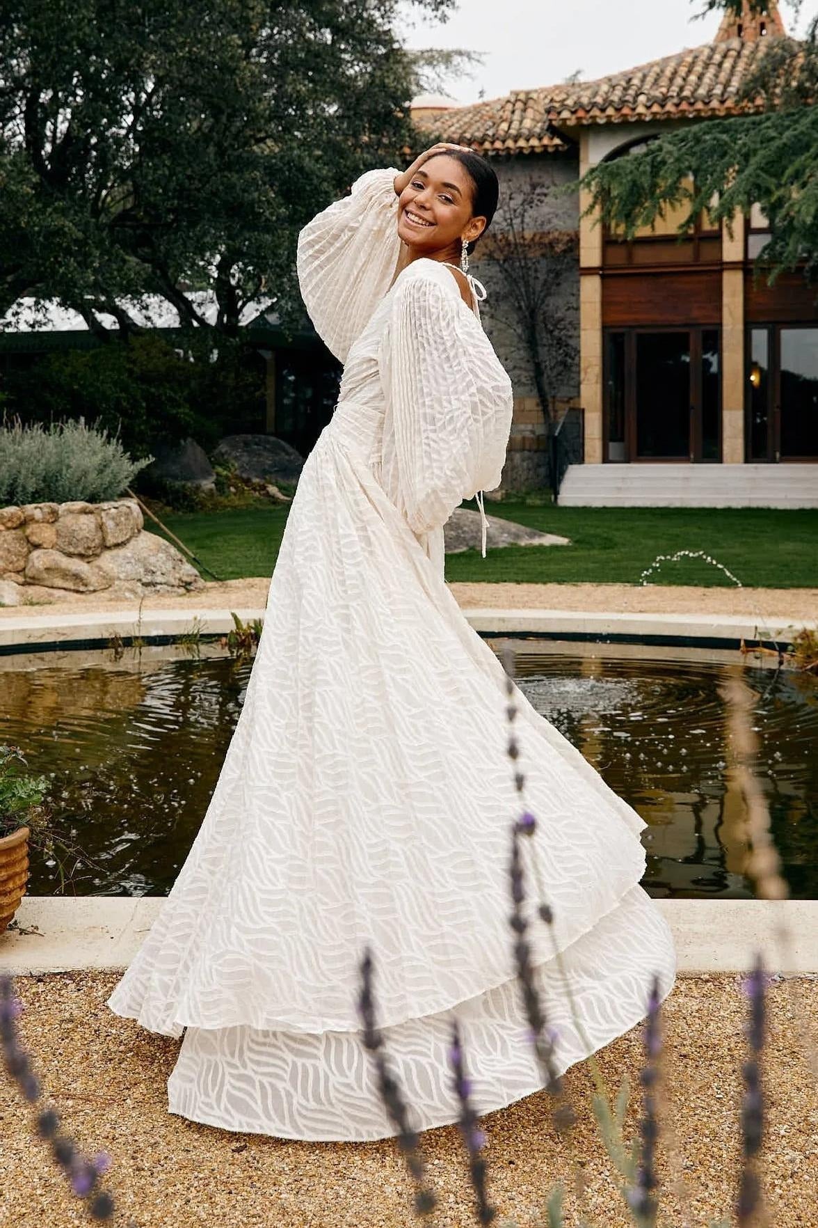 A woman poses in a garden wearing a long-sleeved, off-white maxi dress with a deep V-neck, voluminous bishop sleeves, a ruched waist, and a fully pleated skirt with a subtle wave pattern.
