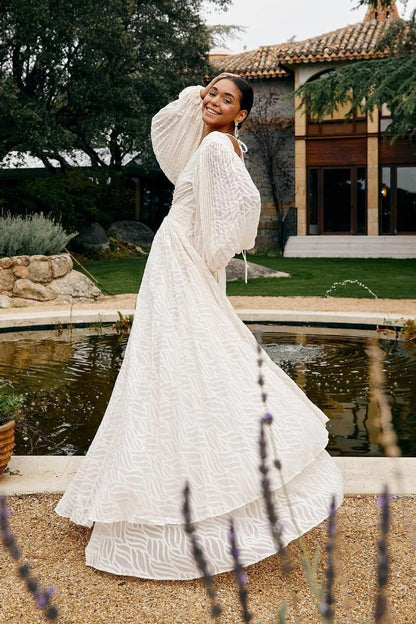 A woman poses in a garden wearing a long-sleeved, off-white maxi dress with a deep V-neck, voluminous bishop sleeves, a ruched waist, and a fully pleated skirt with a subtle wave pattern.