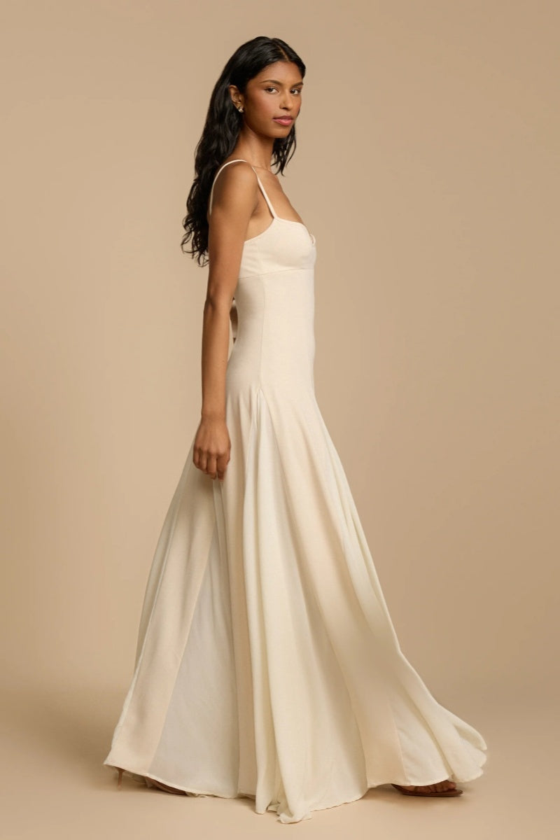 Full-length studio shot of a woman wearing a cream-colored A-line maxi dress with spaghetti straps, a sweetheart bustier bodice, and a voluminous, flowing skirt spread out at the hem.