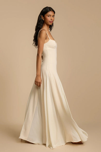 Full-length studio shot of a woman wearing a cream-colored A-line maxi dress with spaghetti straps, a sweetheart bustier bodice, and a voluminous, flowing skirt spread out at the hem.