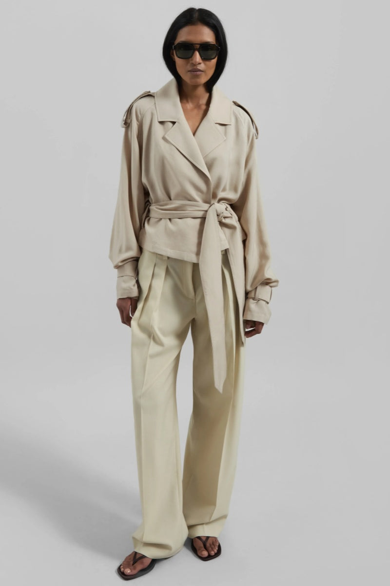 A woman models a light beige, cropped wrap trench jacket featuring wide lapels, shoulder epaulettes, belted cuffs, and a cinched waist tie.