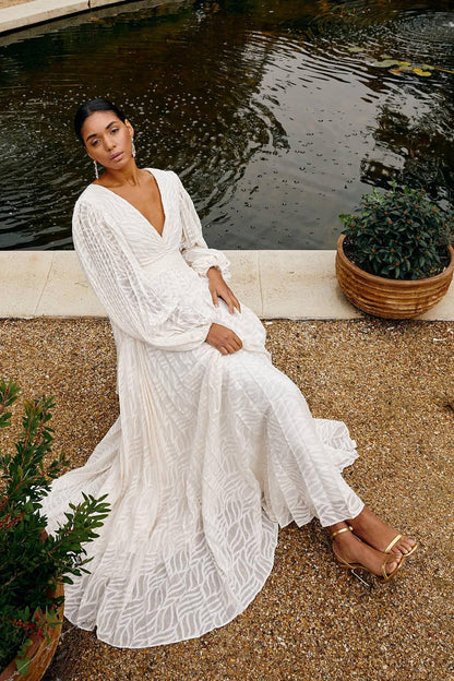 A woman poses in a garden wearing a long-sleeved, off-white maxi dress with a deep V-neck, voluminous bishop sleeves, a ruched waist, and a fully pleated skirt with a subtle wave pattern.