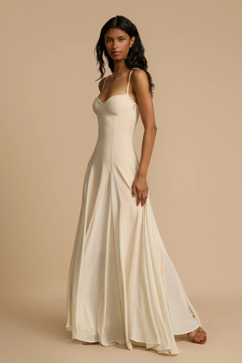 Full-length studio shot of a woman wearing a cream-colored A-line maxi dress with spaghetti straps, a sweetheart bustier bodice, and a voluminous, flowing skirt spread out at the hem.