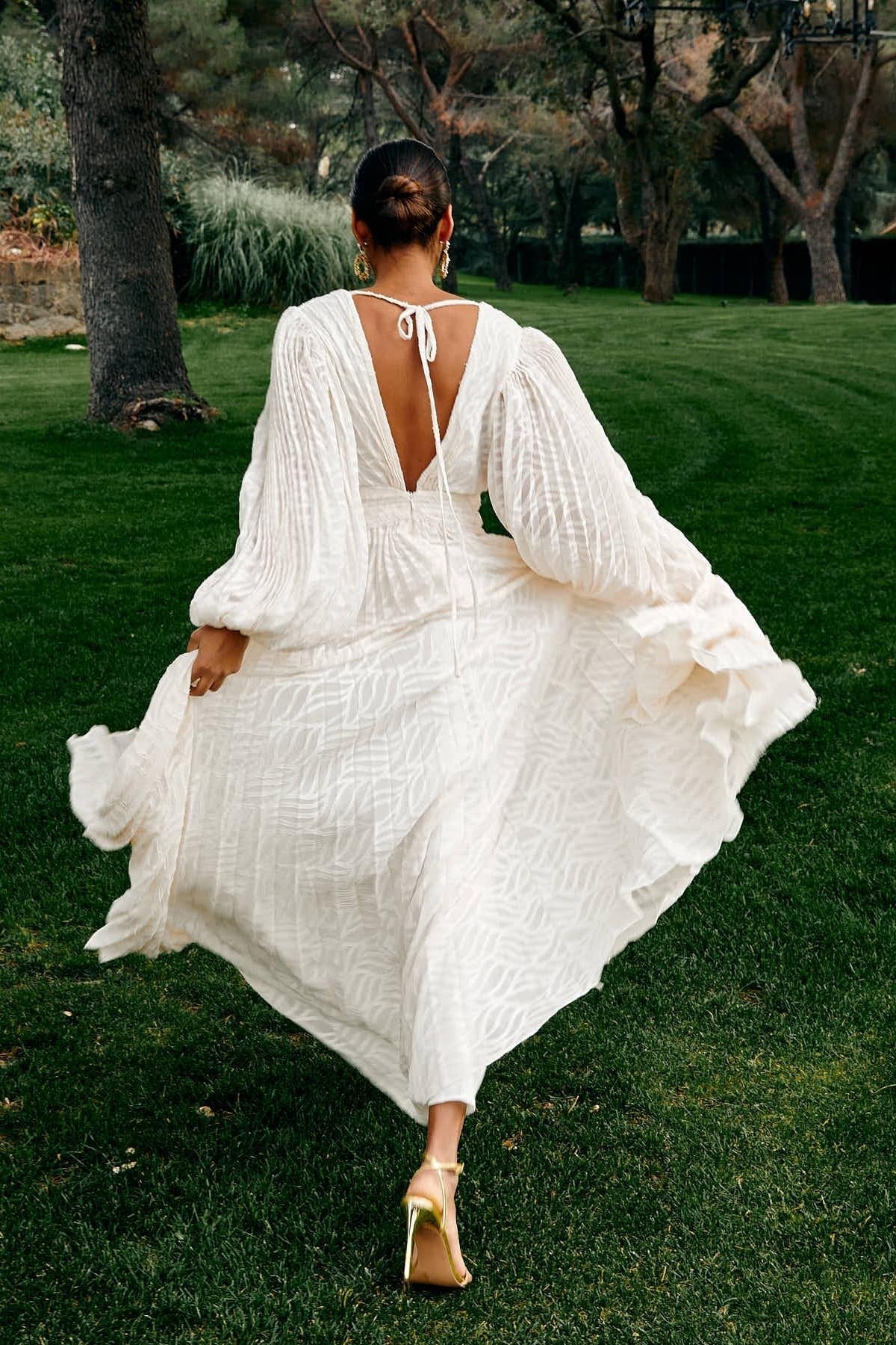 A woman poses in a garden wearing a long-sleeved, off-white maxi dress with a deep V-neck, voluminous bishop sleeves, a ruched waist, and a fully pleated skirt with a subtle wave pattern.