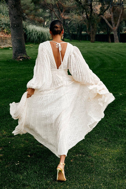 A woman poses in a garden wearing a long-sleeved, off-white maxi dress with a deep V-neck, voluminous bishop sleeves, a ruched waist, and a fully pleated skirt with a subtle wave pattern.