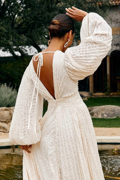 A woman poses in a garden wearing a long-sleeved, off-white maxi dress with a deep V-neck, voluminous bishop sleeves, a ruched waist, and a fully pleated skirt with a subtle wave pattern.
