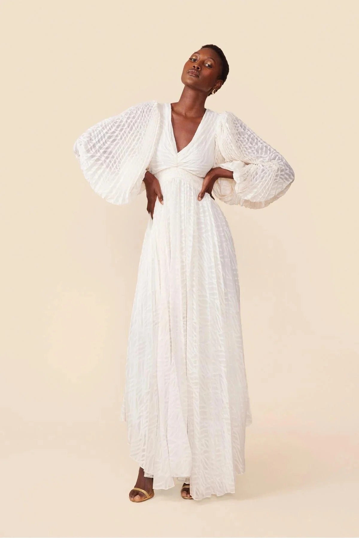 A woman poses in a garden wearing a long-sleeved, off-white maxi dress with a deep V-neck, voluminous bishop sleeves, a ruched waist, and a fully pleated skirt with a subtle wave pattern.