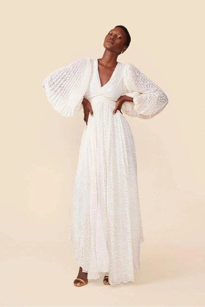 A woman poses in a garden wearing a long-sleeved, off-white maxi dress with a deep V-neck, voluminous bishop sleeves, a ruched waist, and a fully pleated skirt with a subtle wave pattern.