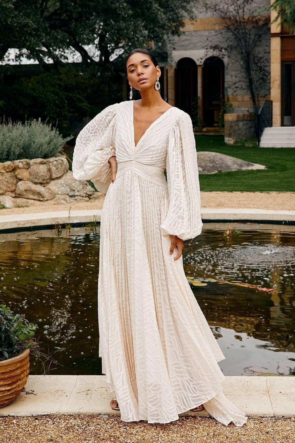 A woman poses in a garden wearing a long-sleeved, off-white maxi dress with a deep V-neck, voluminous bishop sleeves, a ruched waist, and a fully pleated skirt with a subtle wave pattern.