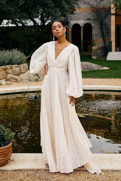 A woman poses in a garden wearing a long-sleeved, off-white maxi dress with a deep V-neck, voluminous bishop sleeves, a ruched waist, and a fully pleated skirt with a subtle wave pattern.