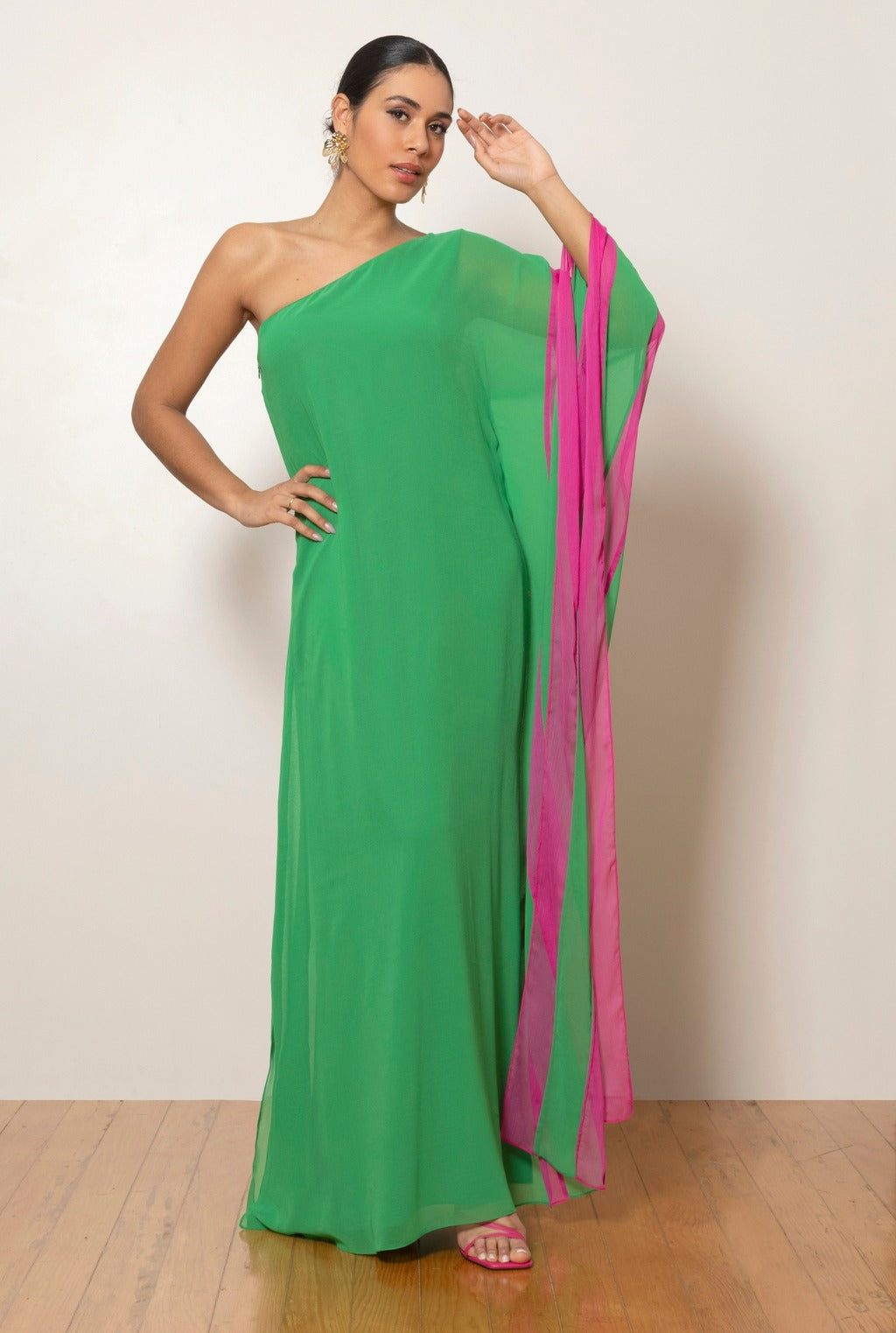 A woman models a vibrant green, one-shoulder, floor-length gown against a white background. The gown has a dramatic, flowing sleeve with a bright pink contrasting interior panel.