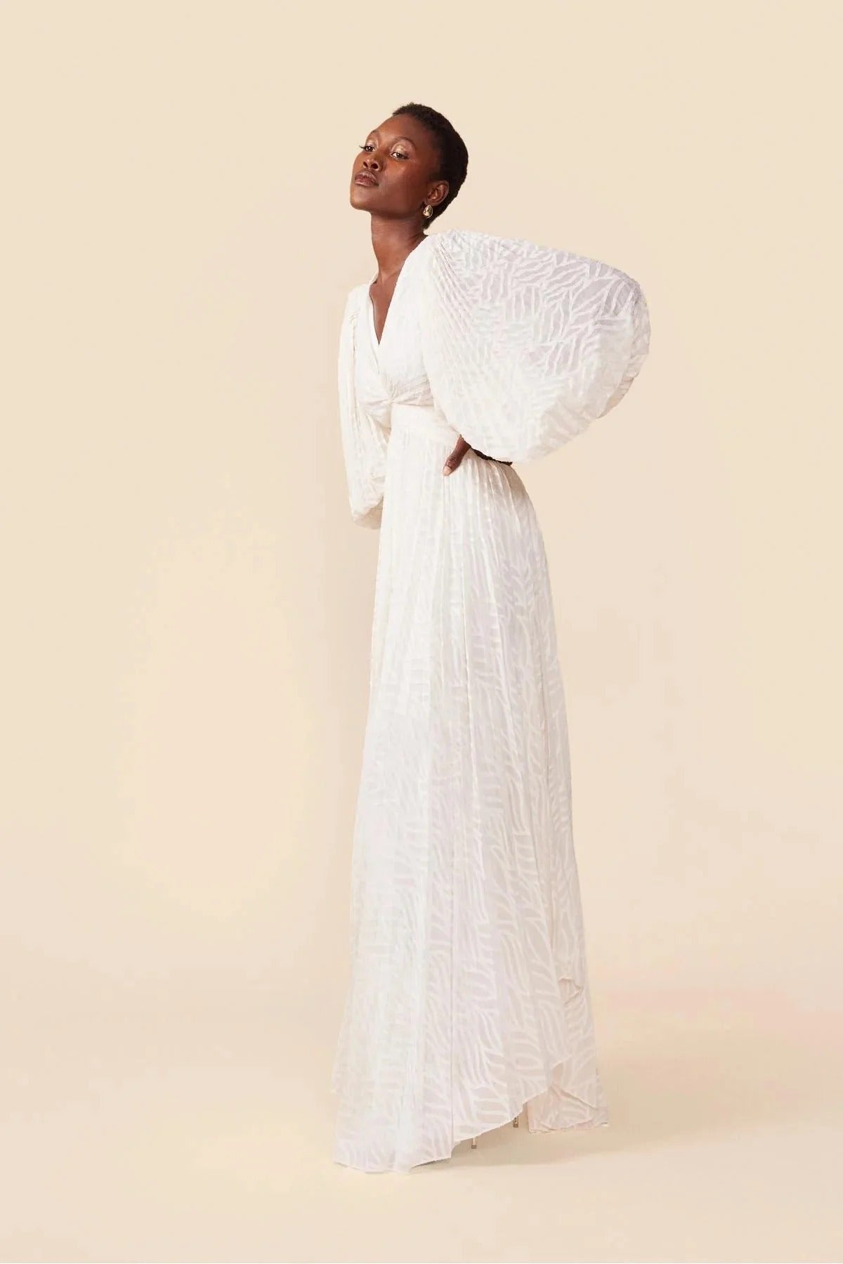 A woman poses in a garden wearing a long-sleeved, off-white maxi dress with a deep V-neck, voluminous bishop sleeves, a ruched waist, and a fully pleated skirt with a subtle wave pattern.
