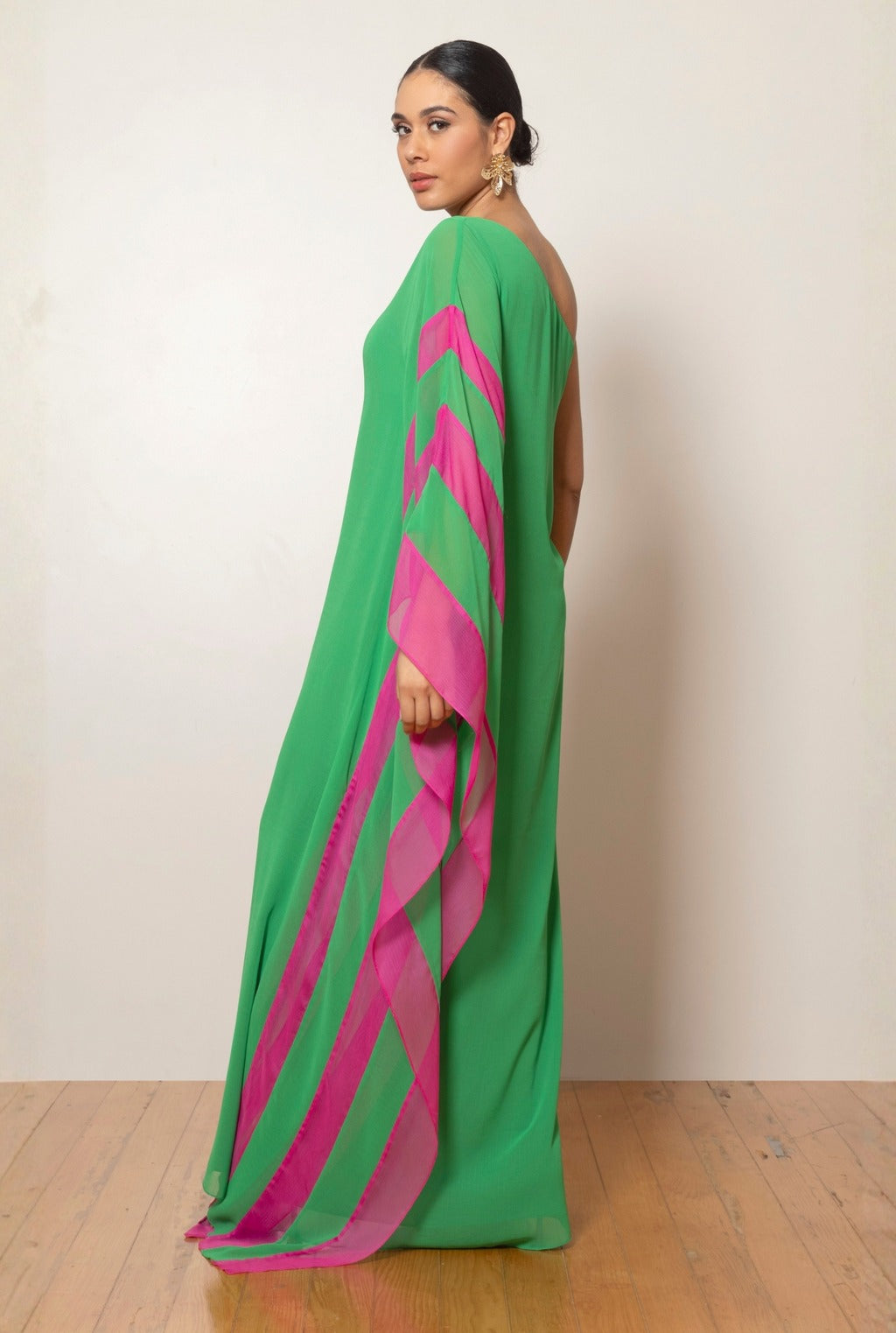 A woman models a vibrant green, one-shoulder, floor-length gown against a white background. The gown has a dramatic, flowing sleeve with a bright pink contrasting interior panel.