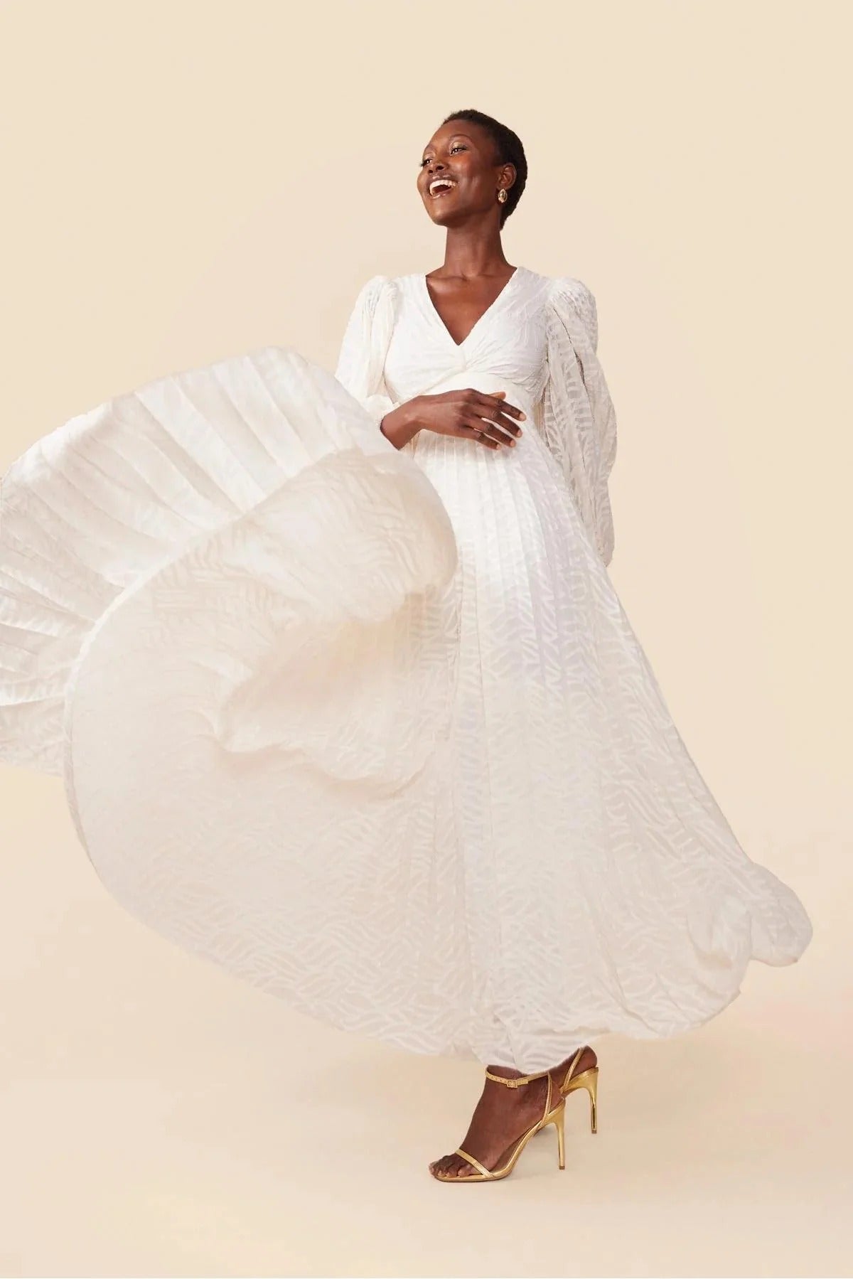 A woman poses in a garden wearing a long-sleeved, off-white maxi dress with a deep V-neck, voluminous bishop sleeves, a ruched waist, and a fully pleated skirt with a subtle wave pattern.