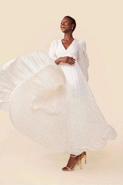 A woman poses in a garden wearing a long-sleeved, off-white maxi dress with a deep V-neck, voluminous bishop sleeves, a ruched waist, and a fully pleated skirt with a subtle wave pattern.