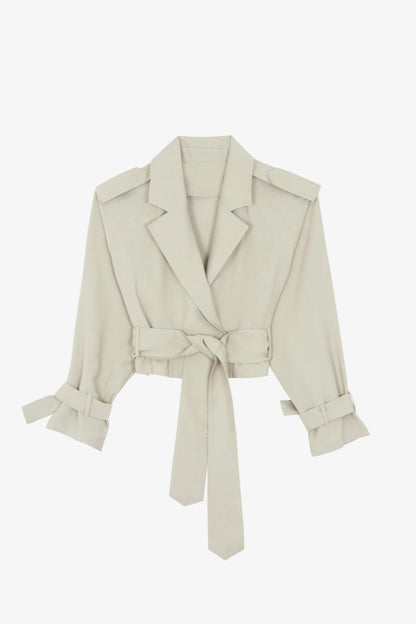 Cement - A woman models a light beige, cropped wrap trench jacket featuring wide lapels, shoulder epaulettes, belted cuffs, and a cinched waist tie.