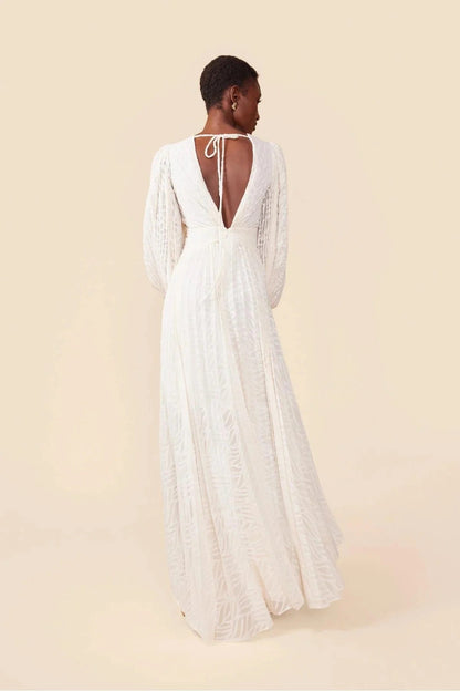 A woman poses in a garden wearing a long-sleeved, off-white maxi dress with a deep V-neck, voluminous bishop sleeves, a ruched waist, and a fully pleated skirt with a subtle wave pattern.