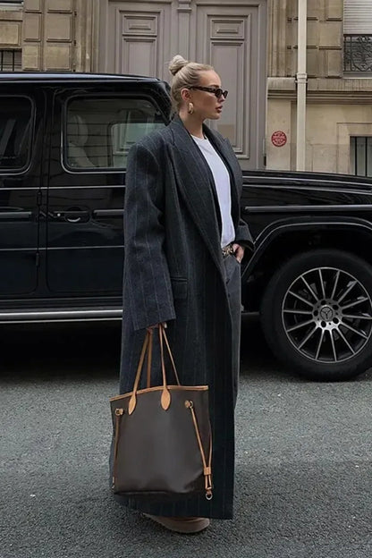 Grey - A woman walking on a city street wearing a dark grey, oversized maxi-length coat with subtle pinstripes, styled over dark jeans and a grey knit hood.