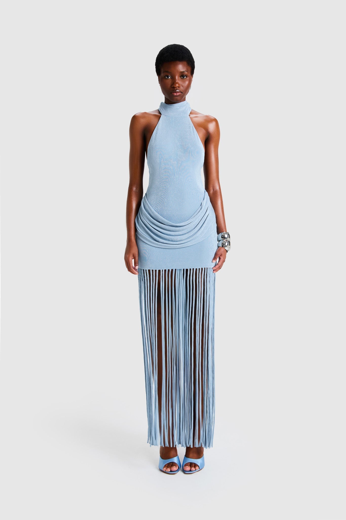 Full-length studio shot of a Black model wearing a sleeveless, high-neck halter dress in a deep red. The dress is form-fitting with ruching at the hips and features a dramatic, floor-length hem made of long, thick corded fringe.