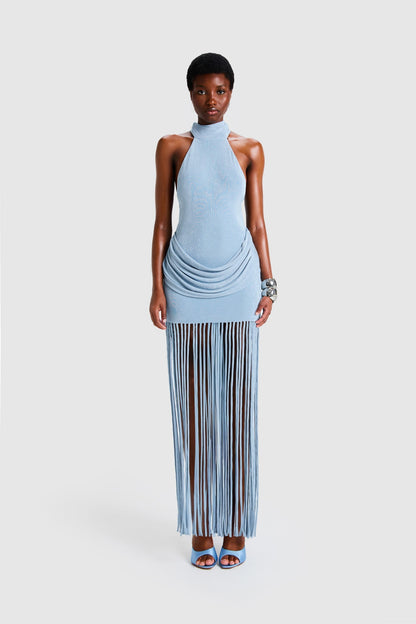 Full-length studio shot of a Black model wearing a sleeveless, high-neck halter dress in a deep red. The dress is form-fitting with ruching at the hips and features a dramatic, floor-length hem made of long, thick corded fringe.
